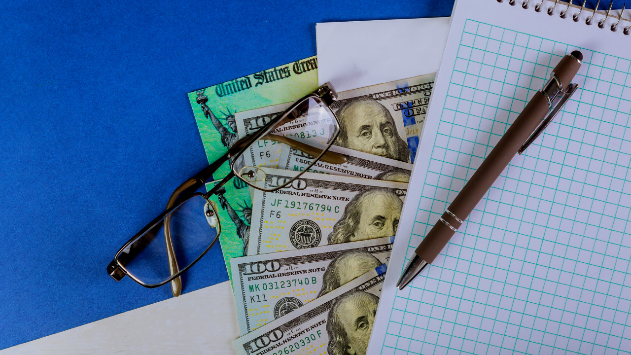 stimulus check with cash and glasses sitting under a pad of paper with a pen