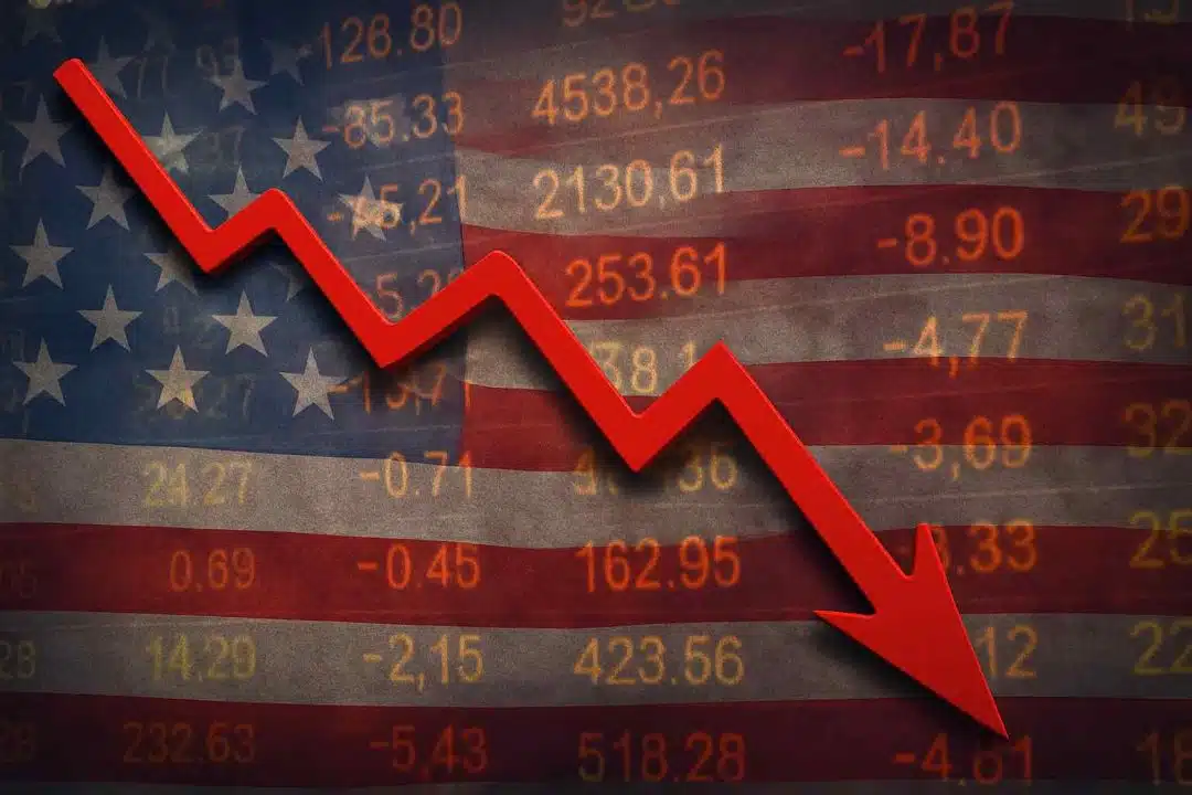 A red downward arrow cuts across an American flag backdrop layered with falling stock market numbers, symbolizing a sharp U.S. economic decline.